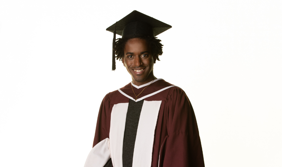 A headshot of Shawn Hercules. He is wearing a ceremonial maroon, black and white graduation gown and cap and is smiling at the camera. He is photographed against a white backdrop.