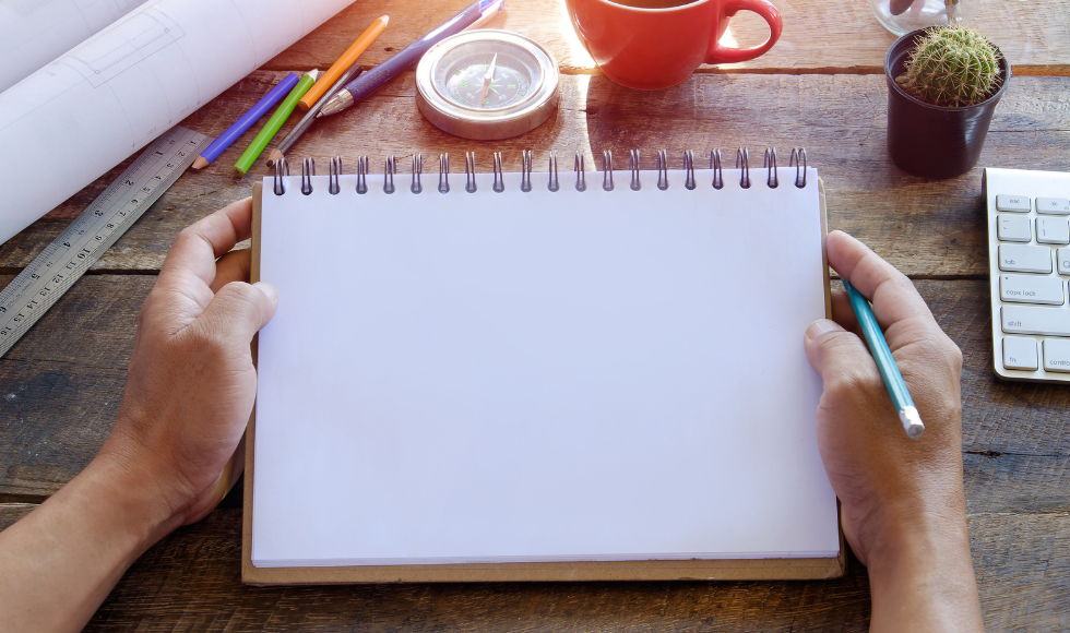 A photo of two hands holding a blank notebook. The hands are resting on a desktop and a keyboard, computer and mug are partially visible. Coloured pencils, a ruler, compass and desk plant are sitting on the desk.