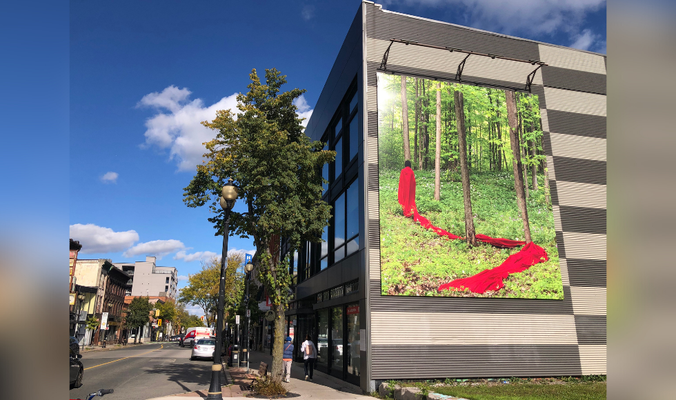 A photo of a large mural on the side of a building on a downtown street. The mural is of a woman wrapped in a long, bright red cloth, standing in a forest. The red material trails behind the woman, snaking along the ground and wrapping around two tress.