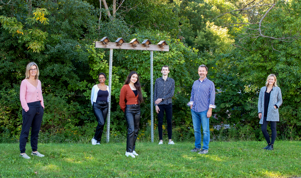 A photo of six people standing outdoors and smiling at the camera. They are standing in front of a pair of posts sticking out of the ground that are holding up a beam of wood. Five bee houses sit on top of the beam of wood.
