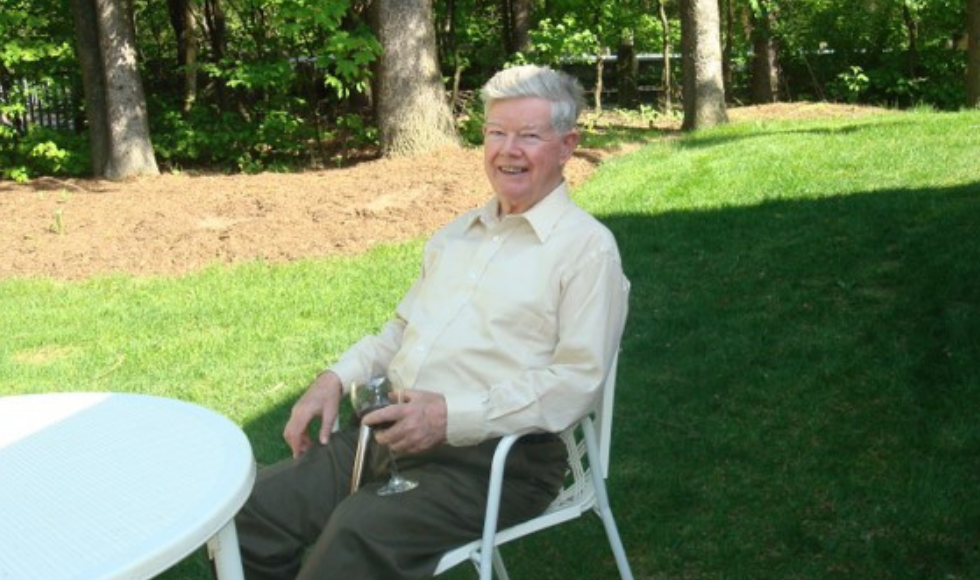 Alan Hitchen smiles for a photo while sitting at a table and holding a wine glass in his left hand. He is outside his Manotick, Ontario home and there are many trees in the background.