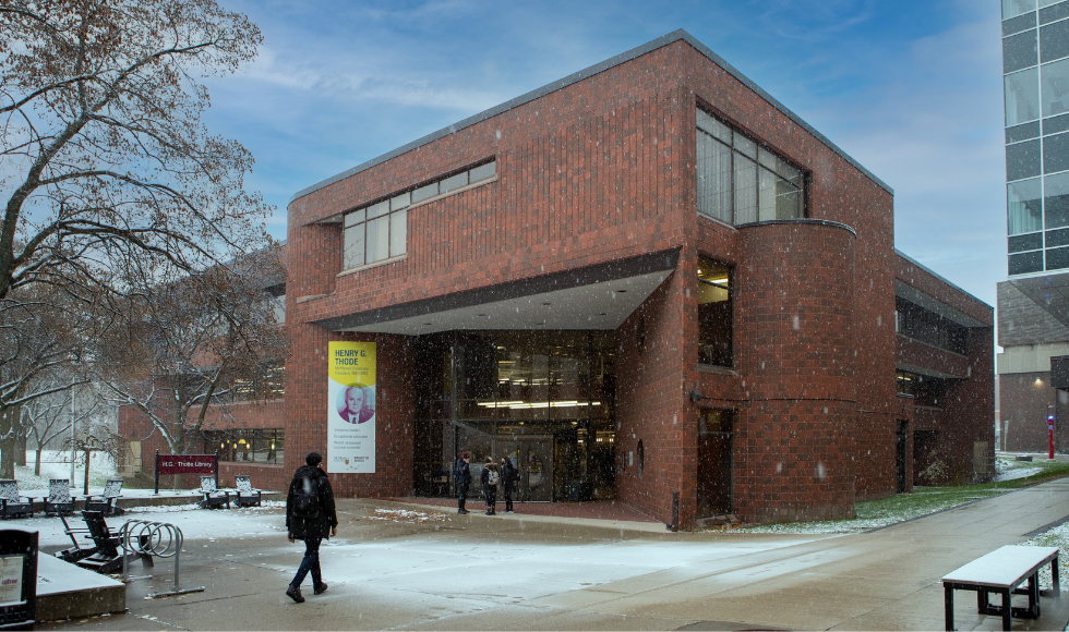 A photo of Thode Library on McMaster University's campus. There is a light dusting of snow on the ground and a tree in front of the building is bare. A person is walking towards the building. Their back is to the camera and their face is not visible.