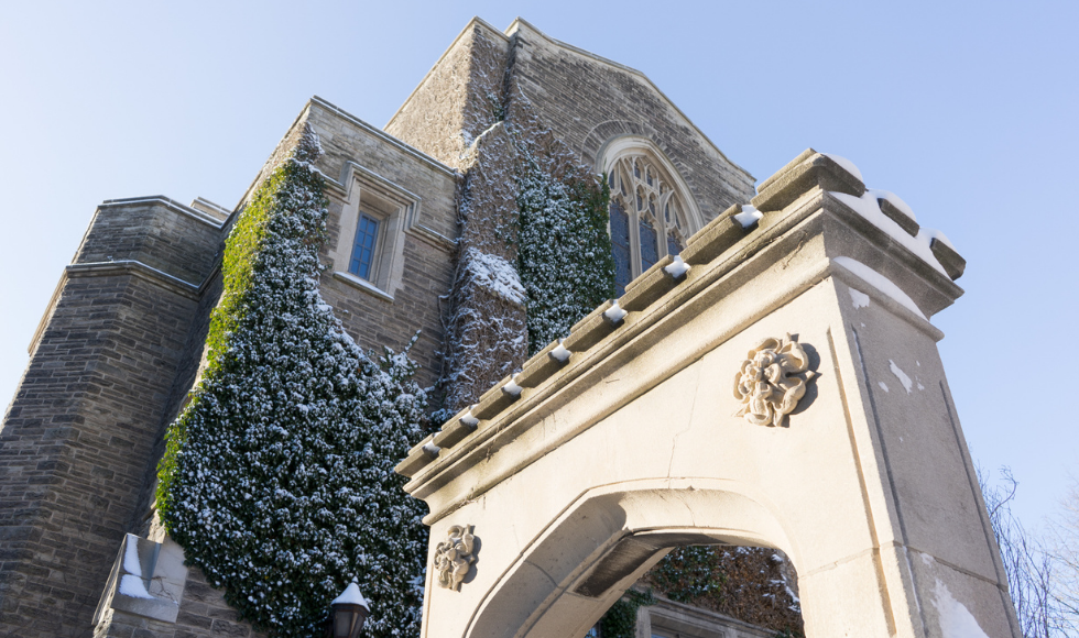 A photo of the Edwards Archway on McMaster's campus. The photo was shot from a low angle looking up at the archway. Edwards Hall can be seen in the background. There is a blue sky in the background.