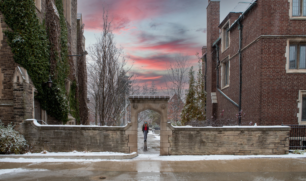 A masked woman walks through archway on McMaster campus. The sky is pink and purple at sunset and patches of snow are on the ground.