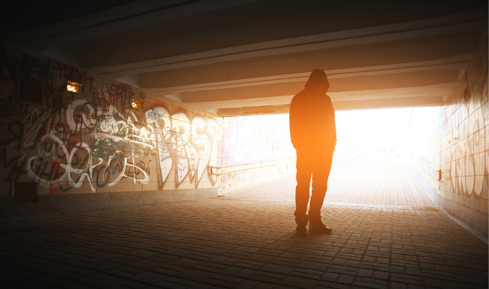 A photo of a person standing with their back to the camera. They are standing under what looks to be a bridge or underpass that is littered with graffiti. A strong beam of light is shining into the area where the person is standing.