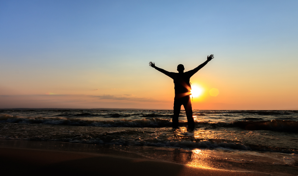 A photo of a man on a beach with his feet in the water looking at a sunset. His arms are outstrectched over his head. His face is not visible.