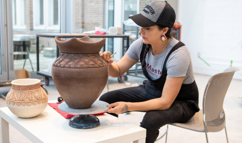 A photo of Santee Smith carving a brown clay pot. She is wearing large earrings, a grey t-shirt, black overalls and a black and grey baseball cap. She is seated on the edge of a chair, working at a table.