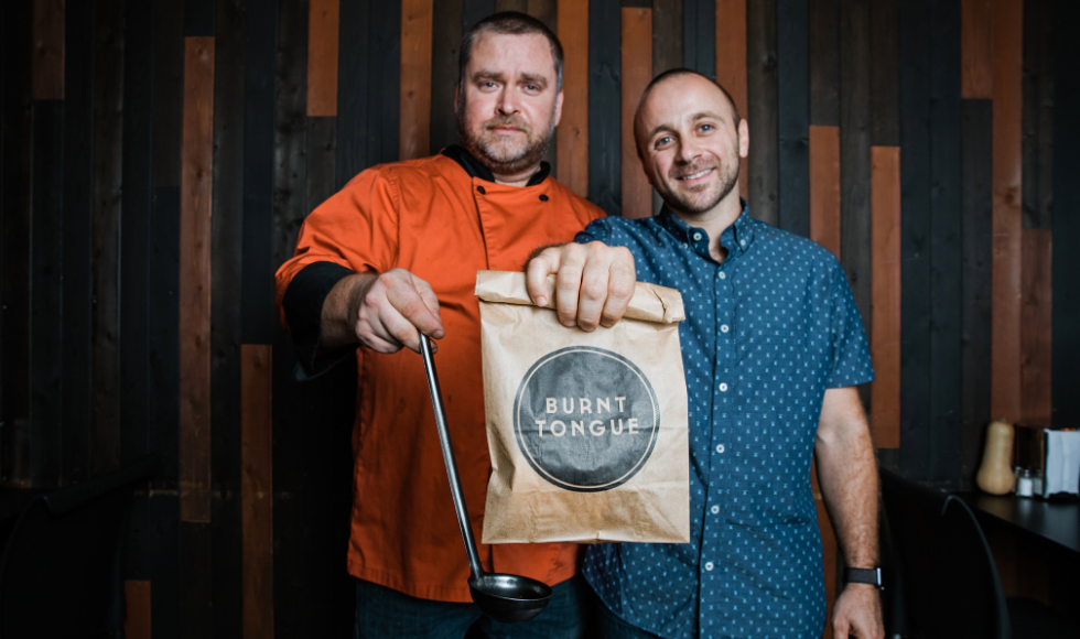 A photo of Leo Tsangarakis and Danny Robinson, the co-founders of the Burnt Tongue chain of restaurants. Tsangarakis is holding a paper bag out in front of him that reads ‘Burnt Tongue,’ and Robinson is holding out a soup ladle in front of him. Tsangarakis is smiling at the camera and wearing a blue patterned shirt. Robinson is in an orange chef’s jacket.