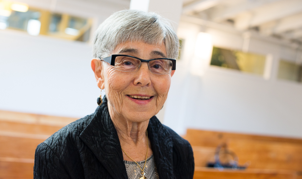 A photo of May Cohen. She is smiling at the camera, wearing glasses, a grey striped shirt and a black jacket. The background is out of focus.