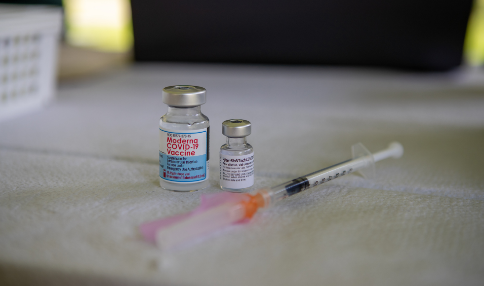 A photo of two vials and a capped needle sitting on a table. One vial is labelled as the Moderna COVID-19 vaccine and the other is labelled a Pfizer COVID-19 vaccine.