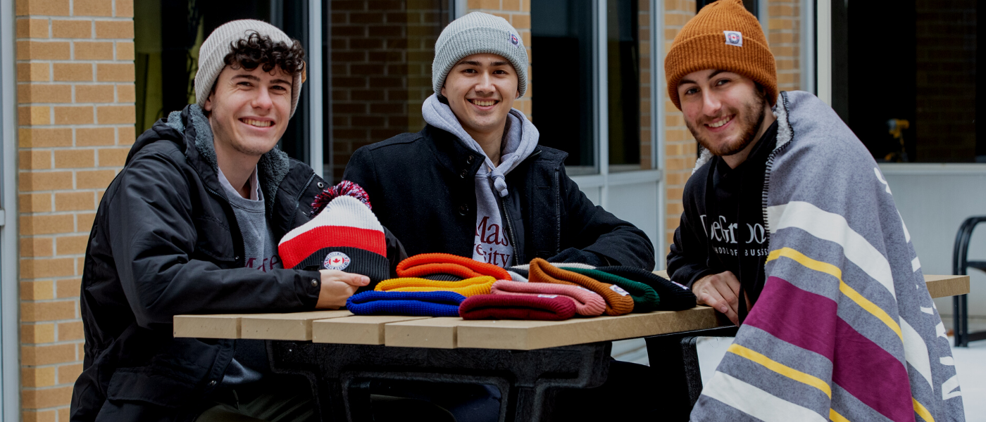 A photo of three McMaster students sitting outside smiling at the camera. They are all wearing toques and one has a McMaster blanket draped over his shoulders.