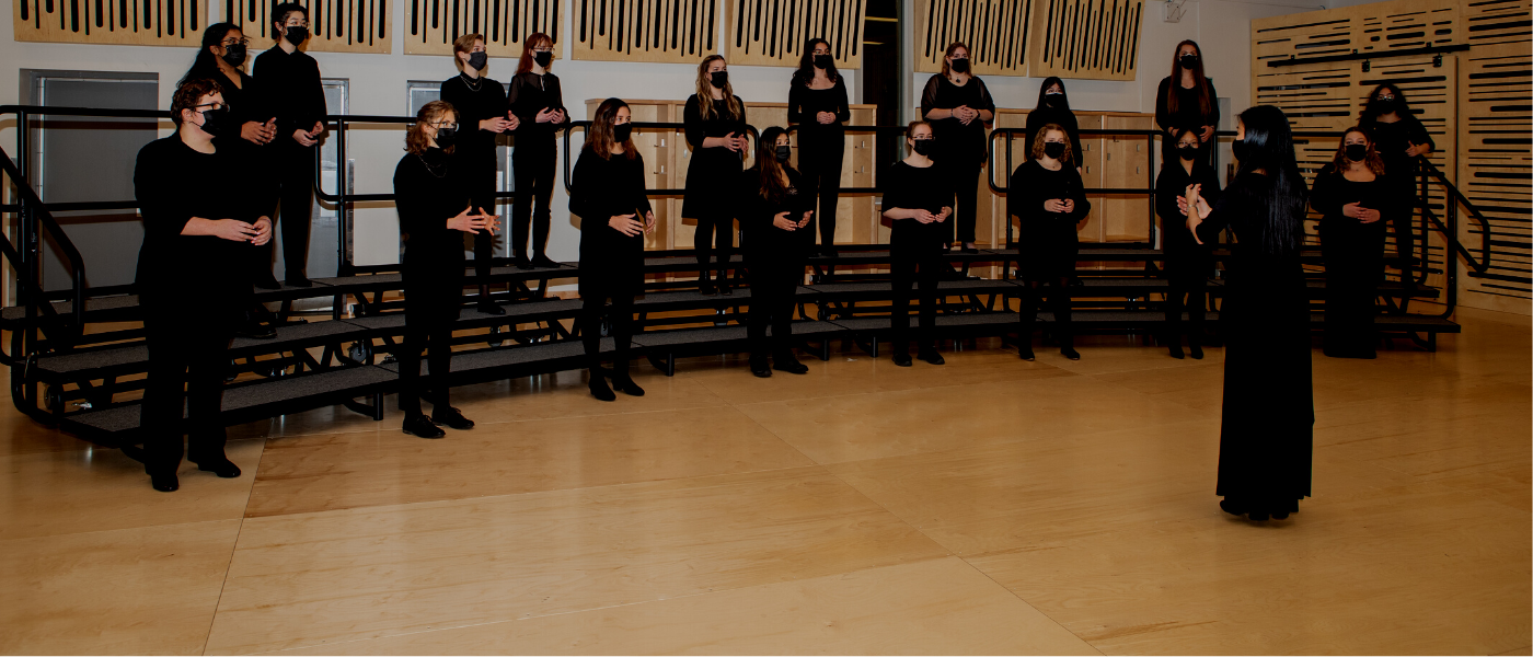 A photo of a choir rehearsing. The choir members are all dressed in black and standing in front of a choir leader.