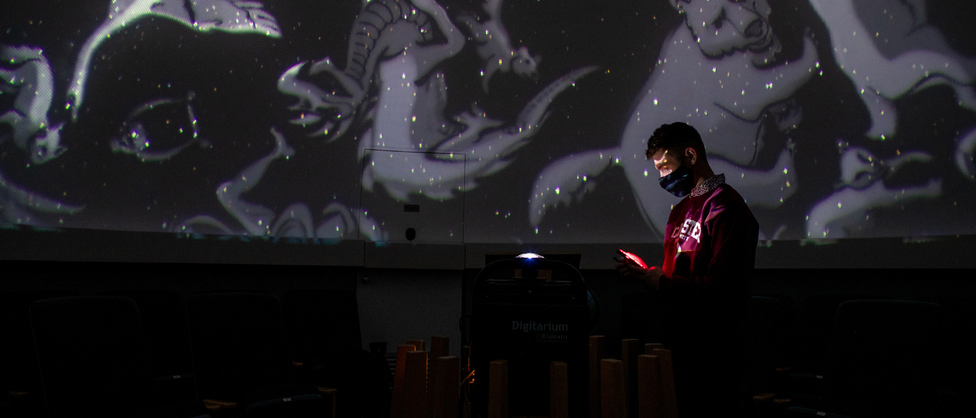 A photo from inside McMaster's Planetarium. A student is standing in front of a projection of constellations. He is wearing a maroon McMaster sweater.