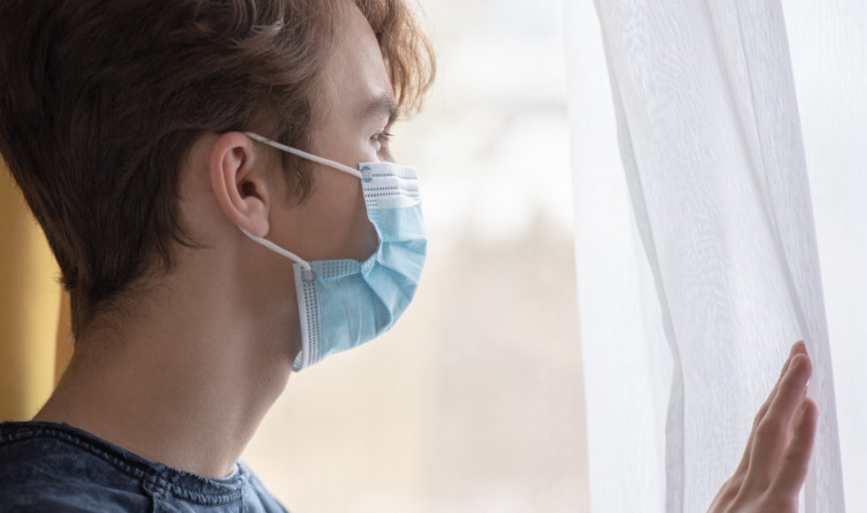 A photo of a man wearing a medical mask and looking out a window. He is pulling back a white curtain.