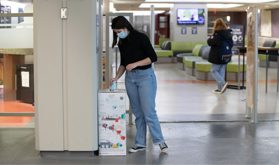A photo of a McMaster student depositing a disposable mask into a recycling bin.