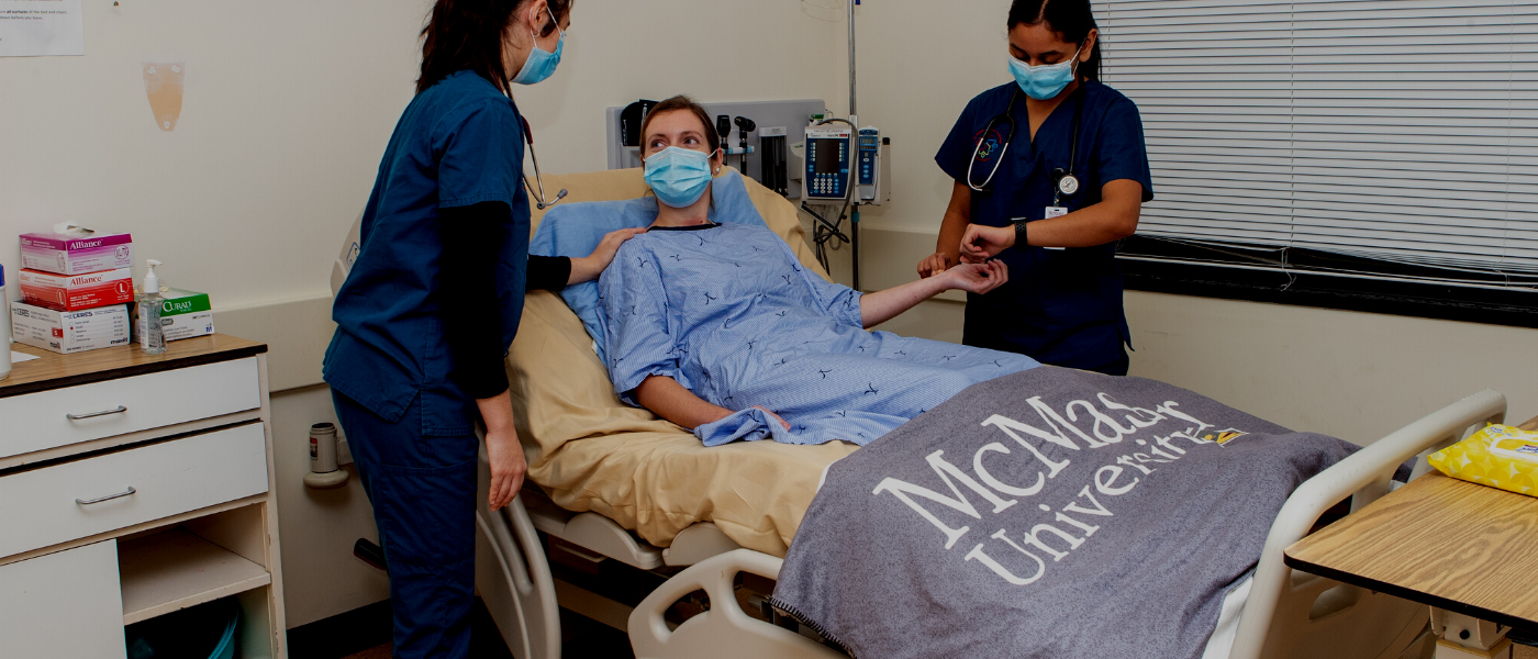 A photo of a patient lying in a hospital bed while two McMaster nursing students tend to them.