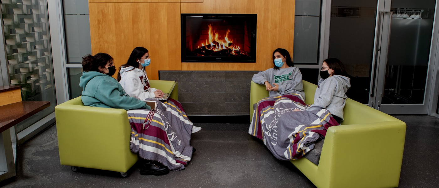 A photo of four McMaster students sitting on two couches in front of a virtual fireplace.