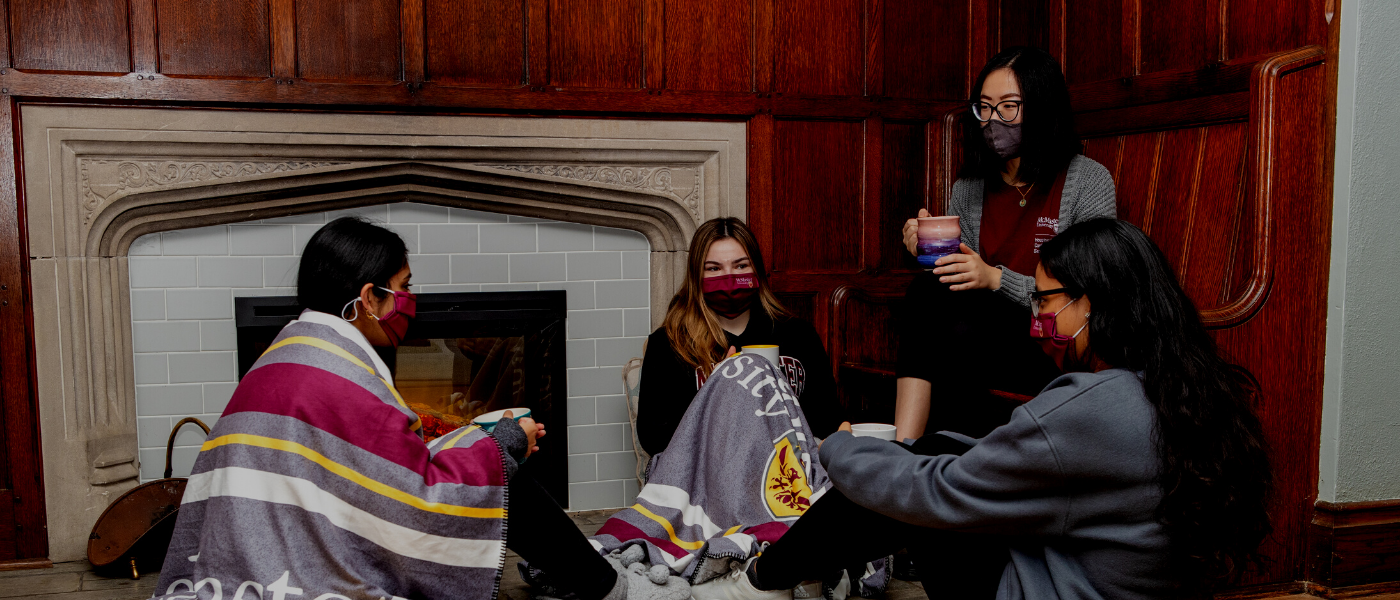 A photo of four McMaster students sitting in front of a fireplace talking. Two have McMaster blankets draped over them.