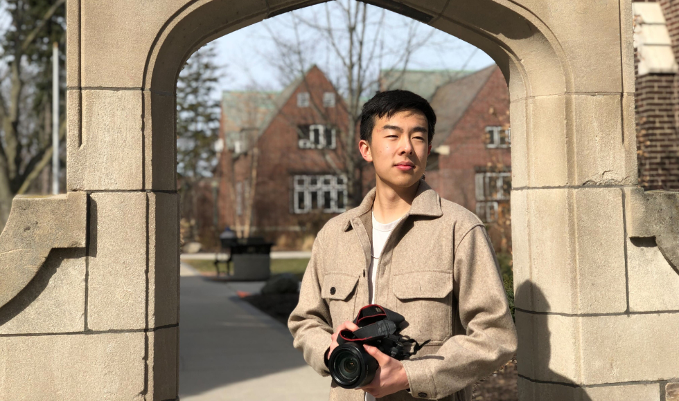 William Liu standing in front of Edwards Arch on McMaster's campus holding a camera