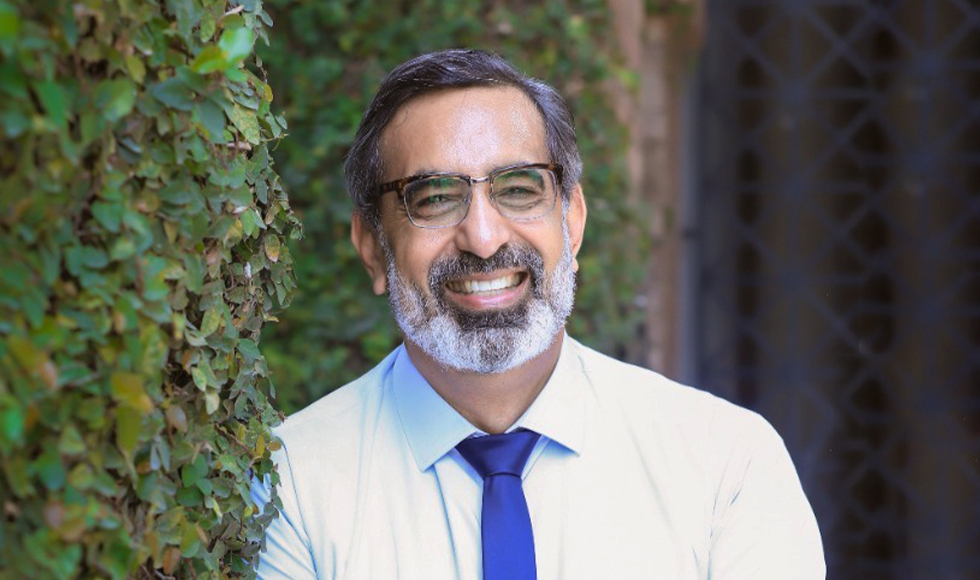 Arshad Ahmad smiling at the camera. He is wearing a white dress shirt and a blue tie and is standing outdoors with greenery behind him.