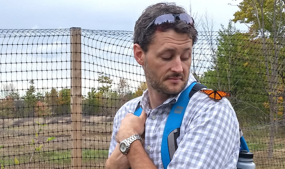 A man wearing a shirt with a collar, monarch butterfly on his shoulder and sunglasses on his head. He is in a field with a fence.