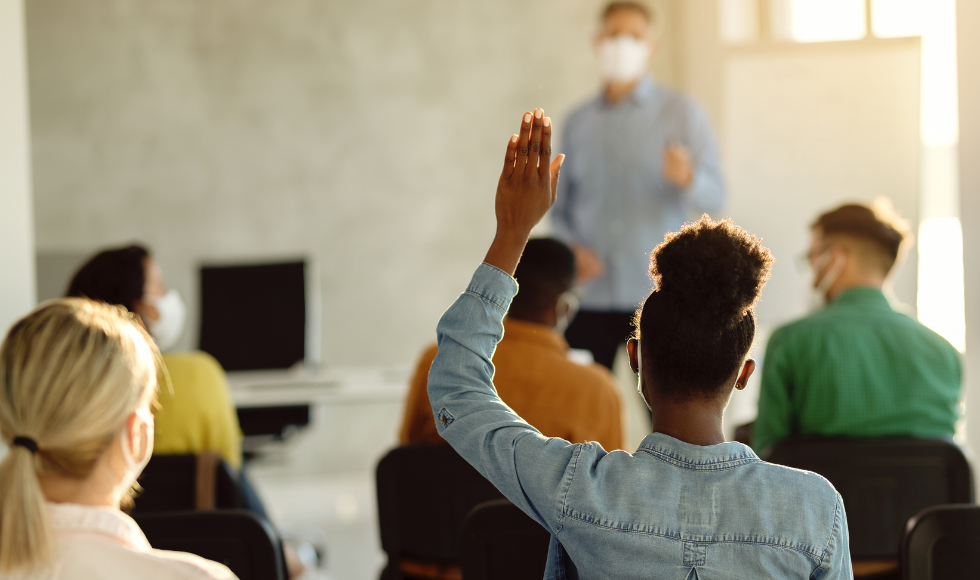 A masked teacher stands at the front of a classroom in front of some seated students. One of the students has their hand up.