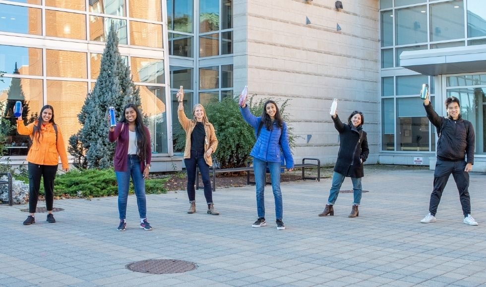 Six people standing in front of a building. They are all holding up a reusable water bottle in their right hands.