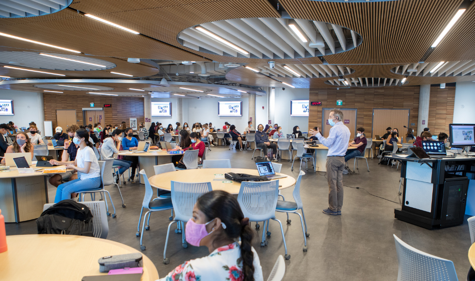 A prof stands in the middle of a room surrounded by students sitting at round tables.