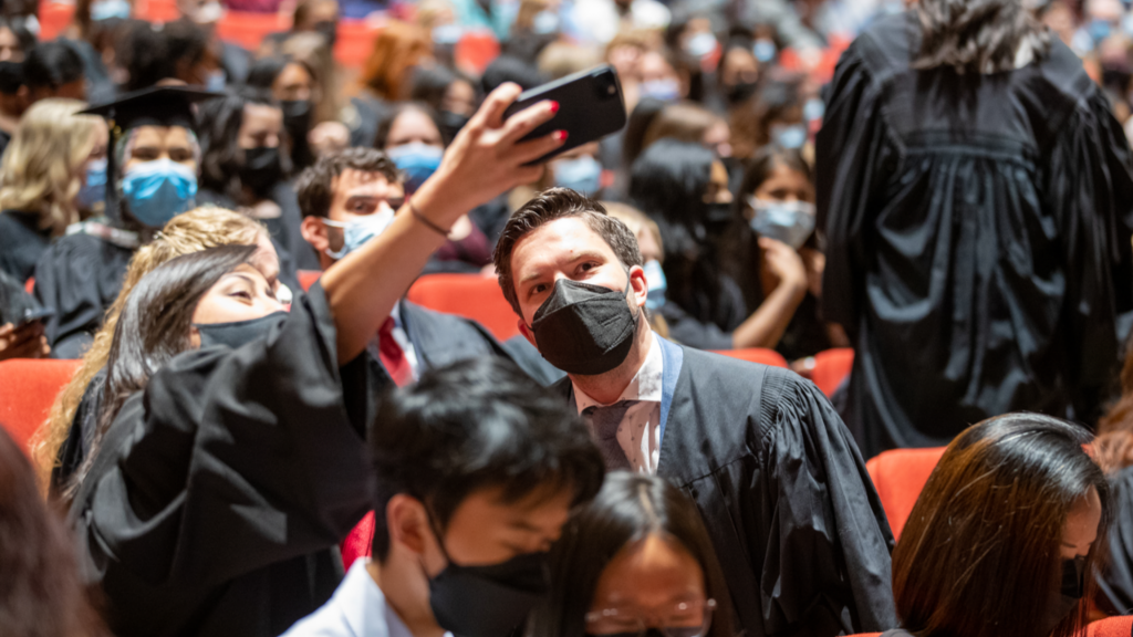 Two students posing for a selfie while sitting in a crowd of students in graduation gowns