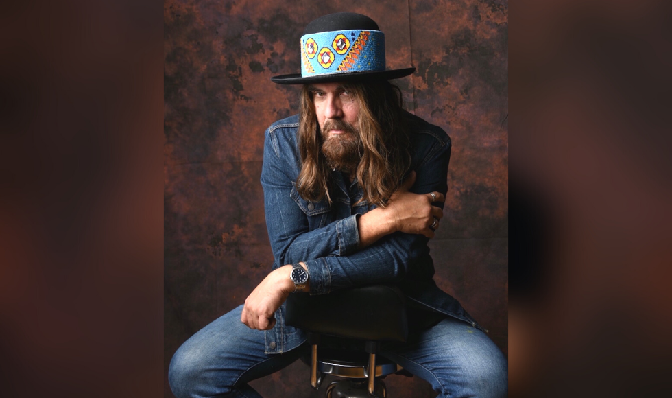 Tom Wilson sitting backward in a chair, wearing denim clothes and a hat and half-smiling