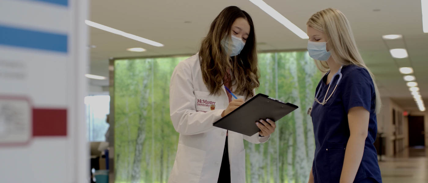 A woman in a white lab coat writes on a clipboard while a woman in blue nursing scrubs looks on