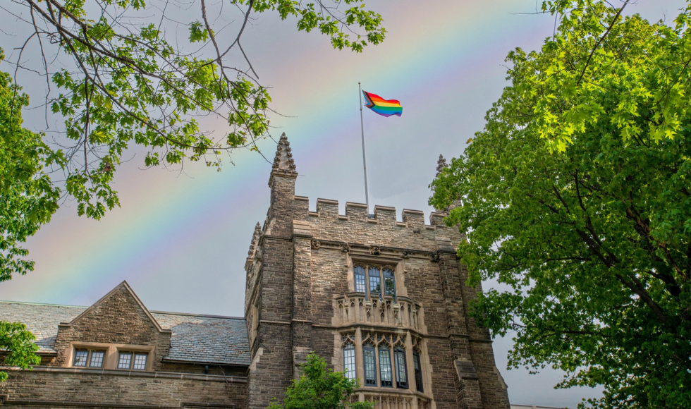 A rainbow flag flying on McMaster’s University Hall. There is a rainbow in the sky behind the flag.