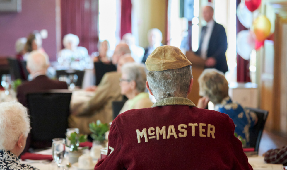 The back of a man's head as he listens to a speech inside McMaster Alumni Memorial Hall. The man is wearing a vintage maroon varsity jacket.