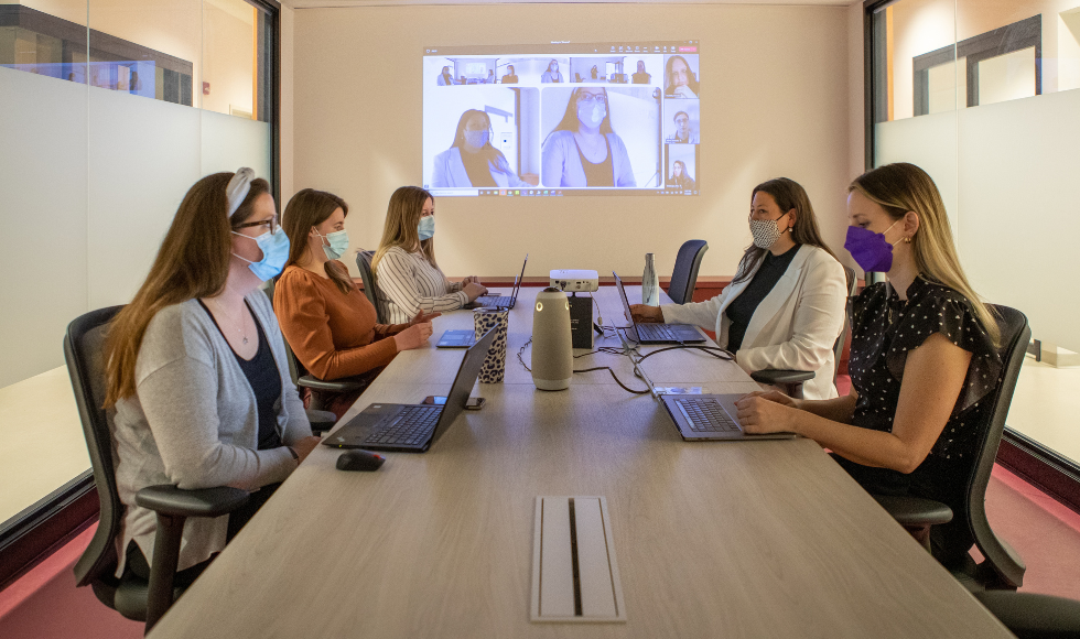 Five masked people in a boardroom sitting around a table with laptops open in front of them. A Microsoft Teams meeting with virtual participants is projected onto the wall at the far end of the room.