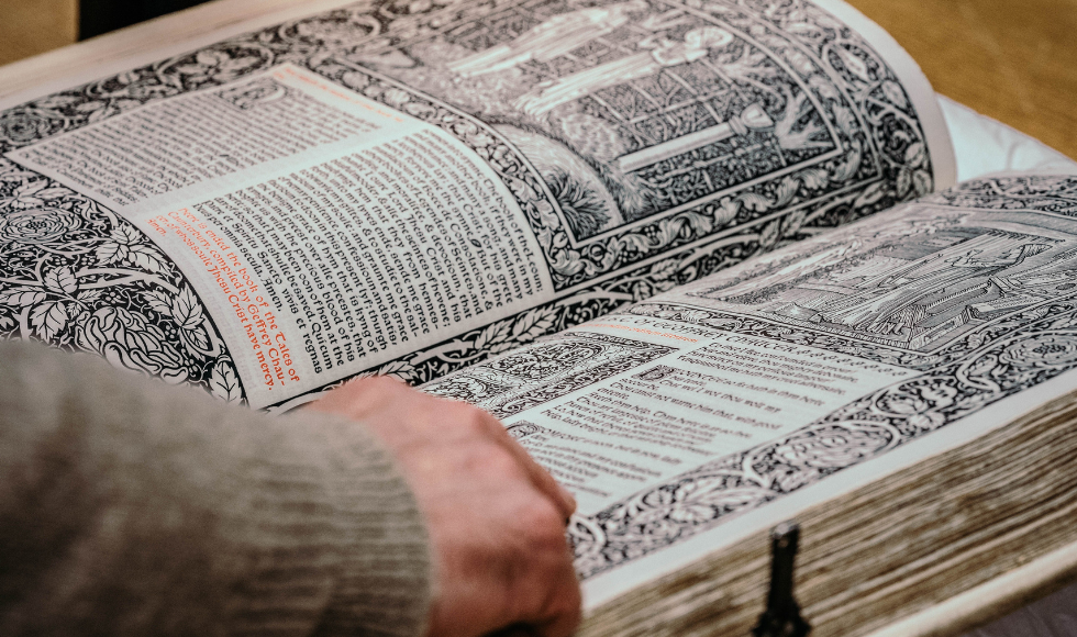 A tight shot of a hand and open ancient book that has ornate illustrations and text on its pages.