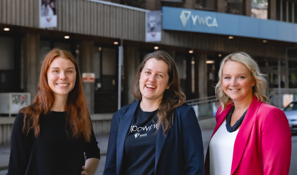 Three women standing and smiling at the camera. Behind them, out of focus, is the exterior of Hamilton’s YWCA.