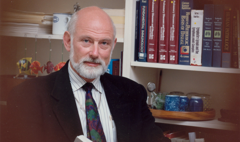John Bienenstock sitting in front of a bookshelf looking directly at the camera. There are medical books, papers and miscellaneous objects on the shelves behind him.