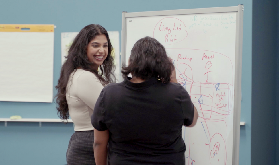 Two students stand at a whiteboard that has words and drawings on it. One student has their head turned and is smiling at the other student whose back is turned to the camera.