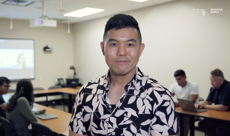 Michael Wong, assistant professor of Psychiatry and Behavioural Neurosciences, standing inside a classroom looking directly at the camera. Behind him there are students seated around tables and a Zoom call projected onto a screen.