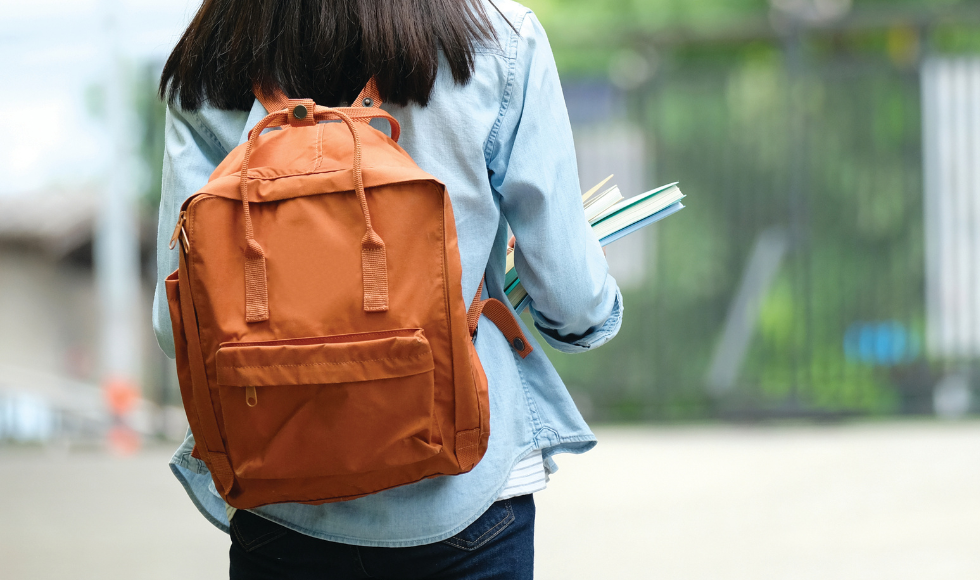 A person carrying books with a backpack on. The photo is taken from behind and the background is out of focus.