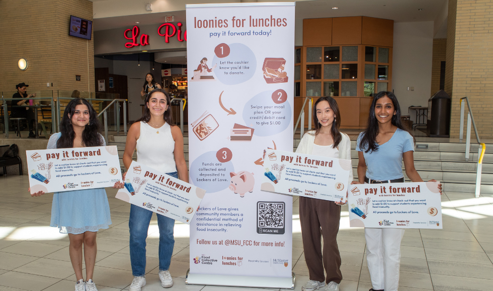 Four students holding up signs stand beside a banner for the Loonies for Lunches banner outside La Piazza on campus.