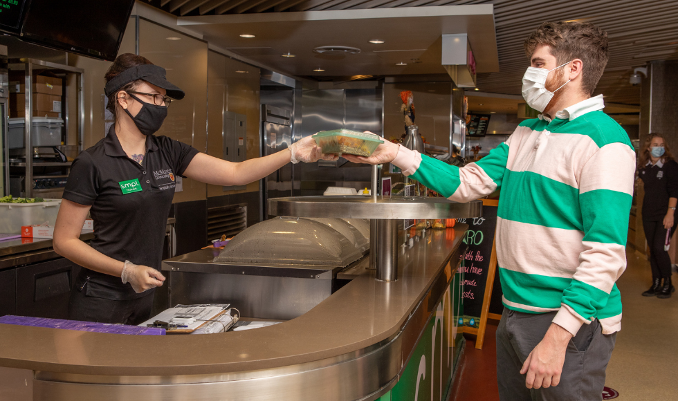 A masked hospitality service worker in Centro hands a student their food, in a compostable container