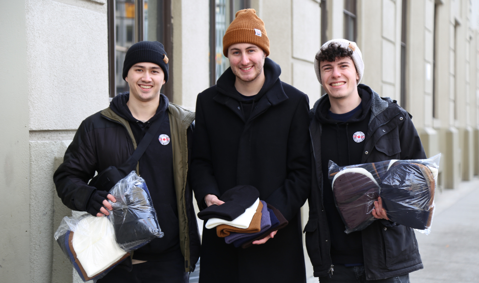 Three people standing outdoors, all wearing toques and smiling at the camera. One is holding a pile of toques, while the other two are holding clear, plastic bags containing toques.