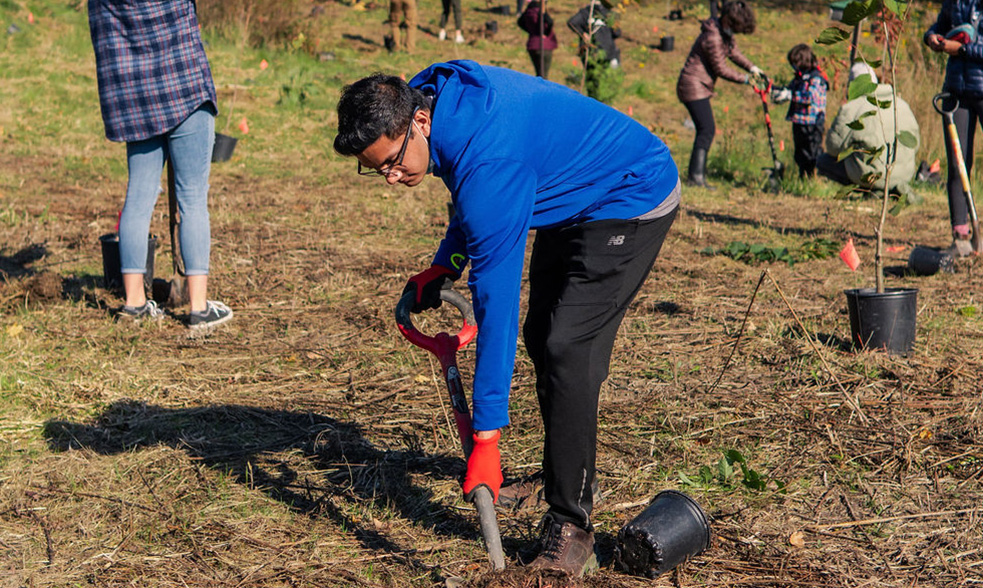 A student in a blue sweater is holding a shovel and digging a hole in a forest. There are people in the background.