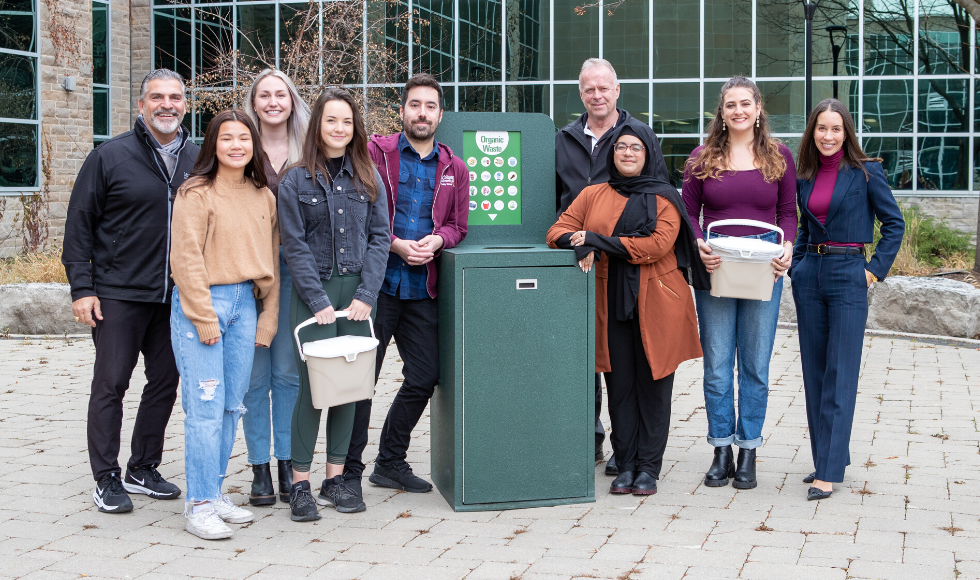 Nine people standing outdoors posing for a photo. In the centre of the photo there is a large, green compost bin. Two of the people are holding smaller, brown and white plastic compost bins.