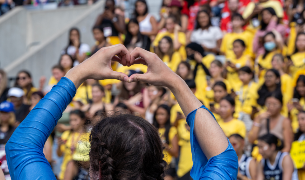 A crowd of people in a stadium out of focus. In the foreground, there is a person with their back to the camera and their hands raised over their head in the shape of a heart.