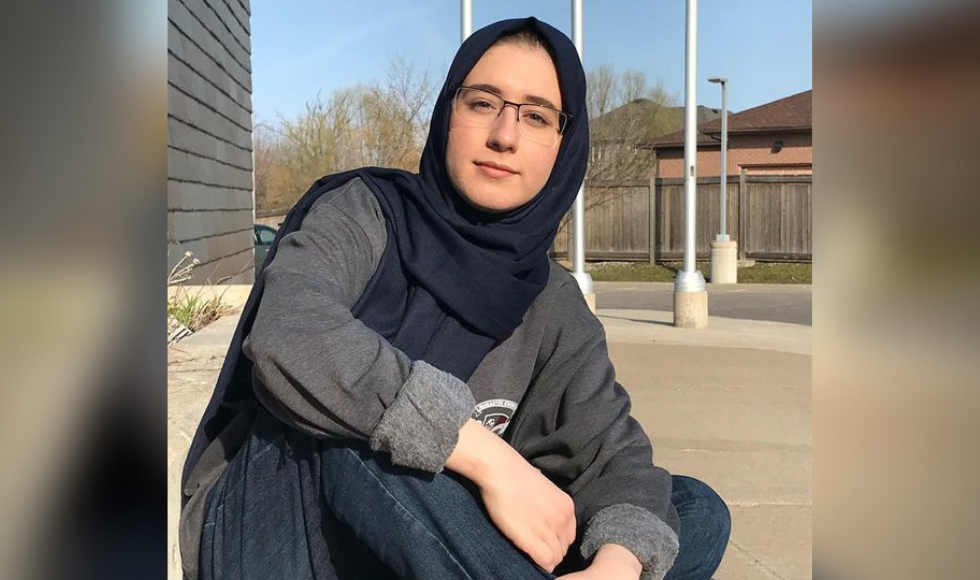 A young woman in a grey sweater wearing a black hijab and glasses looking into the camera.