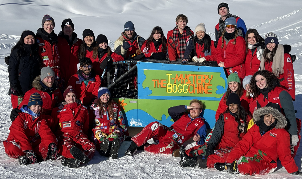 A group of people wearing red snowsuits gathered around a large blue and green five-seater toboggan