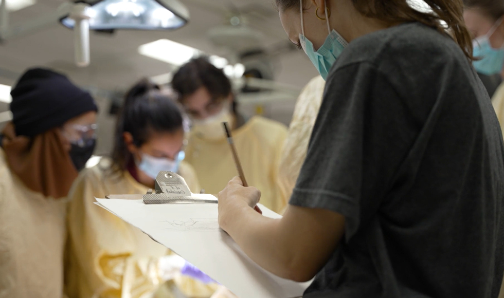 A student sketches in the foreground, while in the background, other students in surgical gear look down at the table [out of frame]
