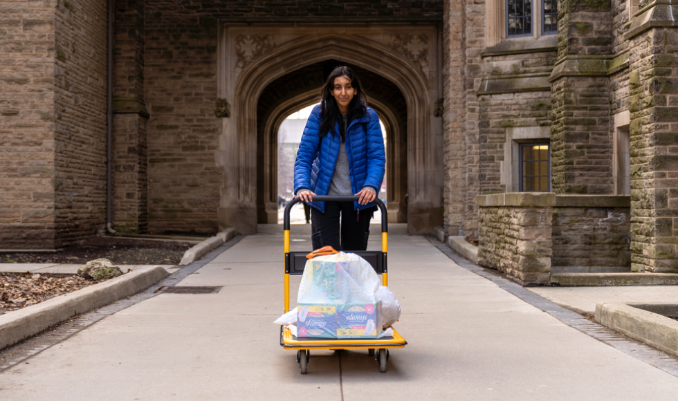 Neha Dhanvanthry with her hands on a push cart that has white plastic bags containing menstrual hygiene products on it.